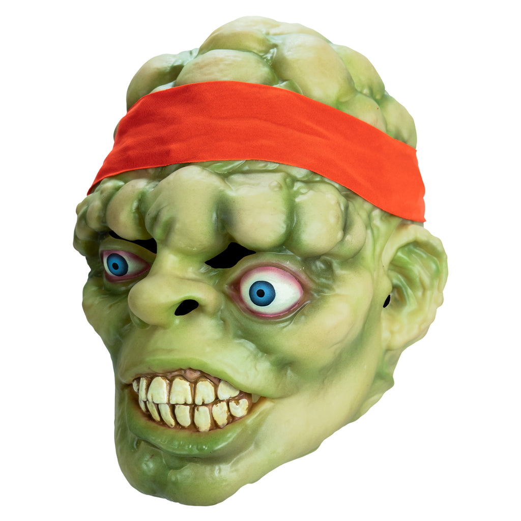 Mask, left side view. Bald head, green lumpy flesh, with red-orange headband tied around forehead. Misaligned, red-rimmed blue eyes, crooked nose. Lips open showing large cracked teeth.