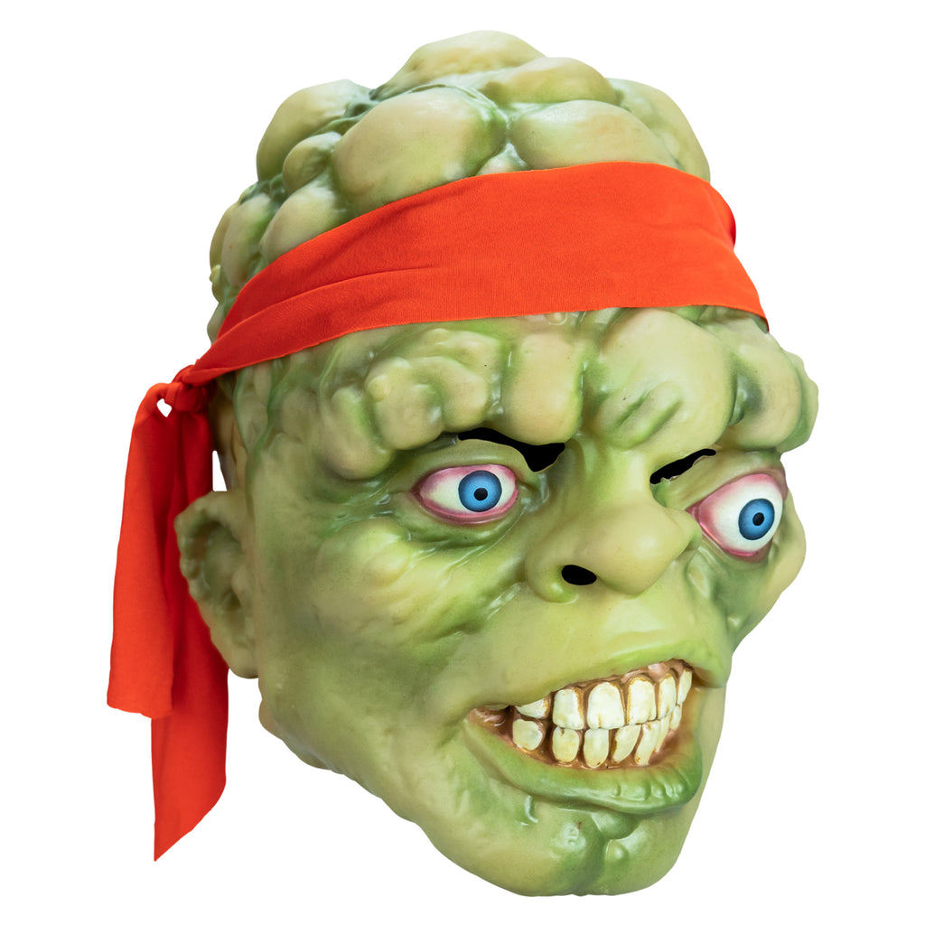 Mask, right side view. Bald head, green lumpy flesh, with red-orange headband tied around forehead. Misaligned, red-rimmed blue eyes, crooked nose. Lips open showing large cracked teeth.