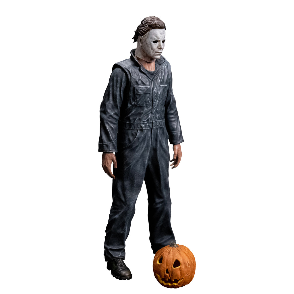 Slight right view, Michael Myers 8 inch action figure. Wearing Halloween (1978) Michael Myers mask, dark coveralls, black boots. arms at sides. Orange jack o' lantern resting on ground in front of the figure