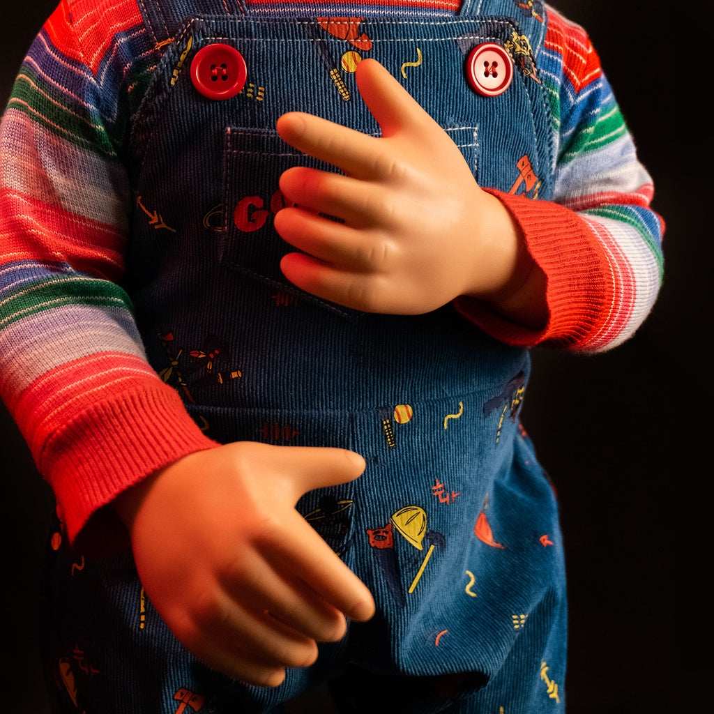 Dramatic red lighting. Close up of Ultimate Chucky doll torso and additional hands, Good Guy Tommy, attached to doll wearing striped shirt under blue overalls. 