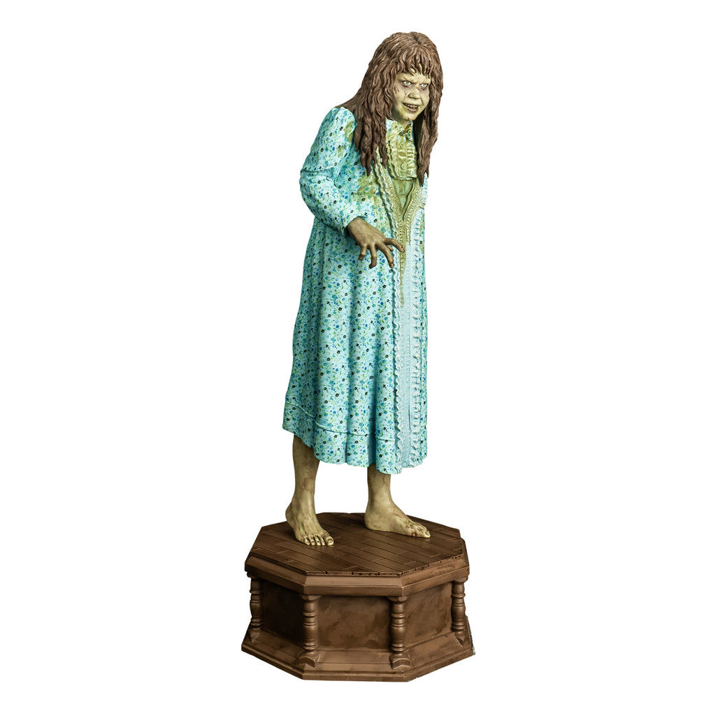 Statue, slight right view. Girl with light brown, wavy hair with bangs. Furrowed brow. red-brown wounds on face. Mouth slightly open, showing upper teeth. Wearing light blue flowered nightgown with large green stain on the front. bare feet. standing on wood-look octagonal base.