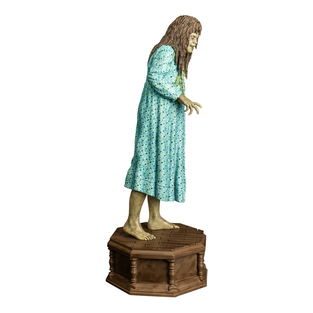 Statue, right side view. Girl with light brown, wavy hair with bangs. Furrowed brow. red-brown wounds on face. Mouth slightly open, showing upper teeth. Wearing light blue flowered nightgown with large green stain on the front. bare feet. standing on wood-look octagonal base.