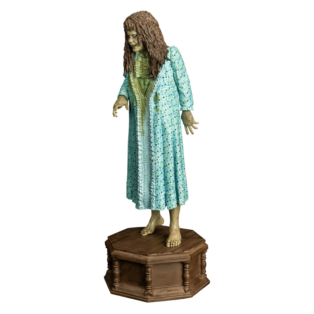 Statue, slight left view. Girl with light brown, wavy hair with bangs. Furrowed brow. red-brown wounds on face. Mouth slightly open, showing upper teeth. Wearing light blue flowered nightgown with large green stain on the front. bare feet. standing on wood-look octagonal base.