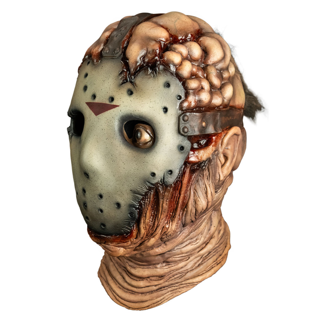 Mask, left side view. lumpy and damaged flesh on top of head, tattered, dirty hockey mask on face, brown straps at top and sides of head from forehead and temples going back. Right empty eye socket, Brown, left eye open, wrinkled and sagging flesh around chin and neck.
