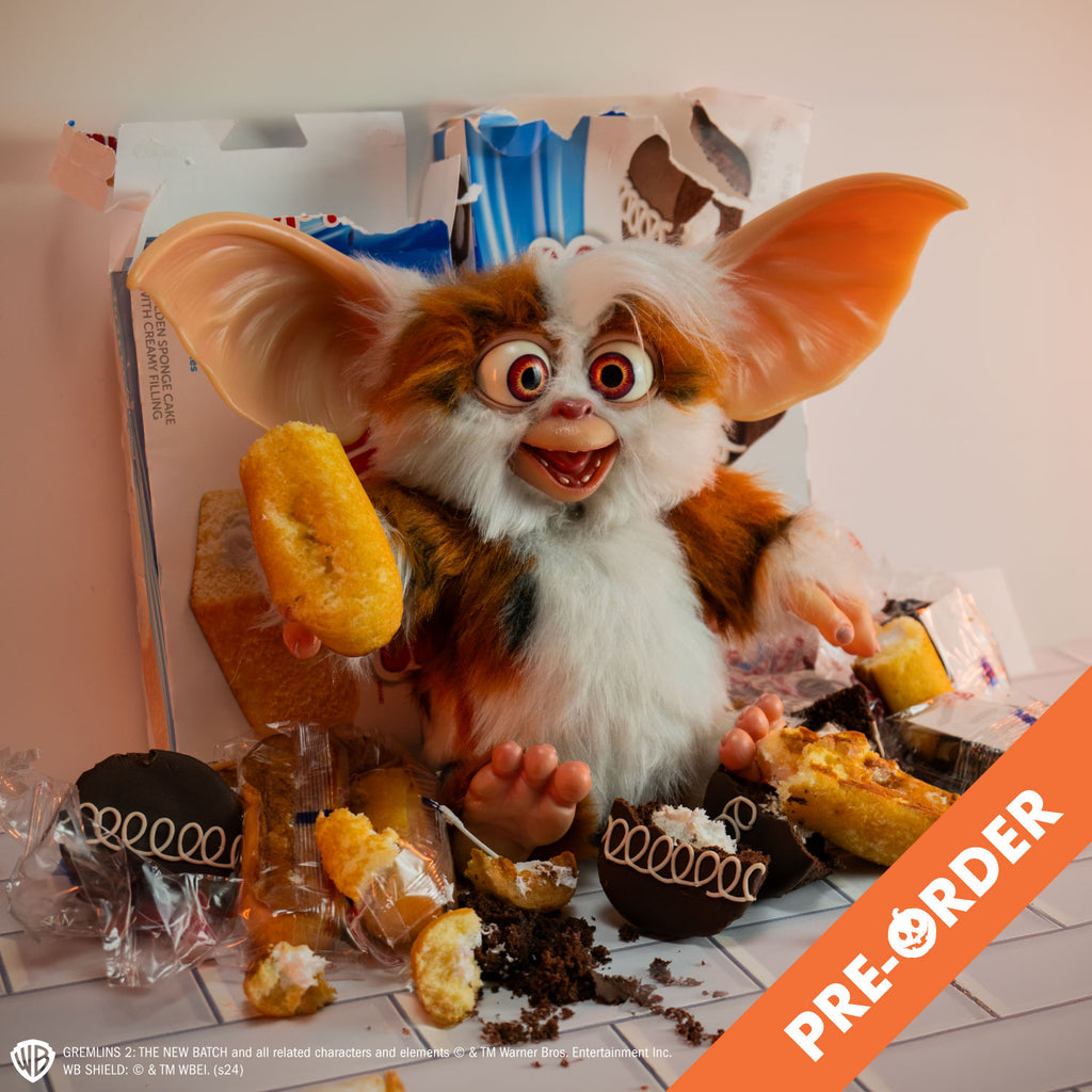 Gremlins 2 prop - Mogwai Daffy, brown and white with some black fur, large crossed eyes, mouth open in a smile, large pointed tan ears. Hairless hands and feet. sitting on a tiled counter surrounded by half-eaten snack cakes, holding one in his right hand.  White text at bottom is licensing information.  Orange diagonal banner at bottom right corner, white text reads pre-order.