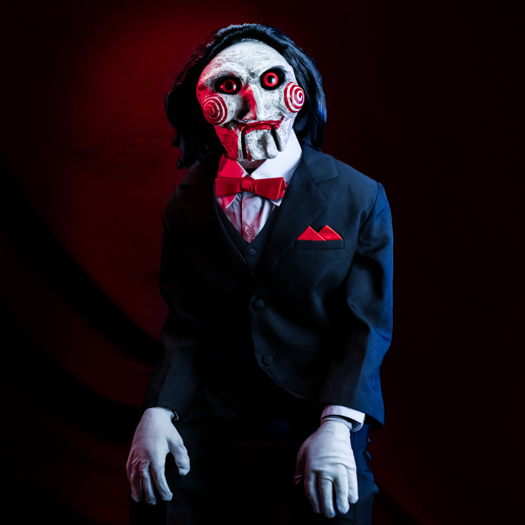 Closeup glamour view, red and black background.  Saw Billy Puppet deluxe prop, balding with black hair, white face, black-rimmed red eyes, red spirals on cheeks, red lips on hinged ventriloquist dummy mouth. Wearing red bowtie, white collared shirt, black vest and suit coat, white gloves, black pants. Sitting with hands on knees.