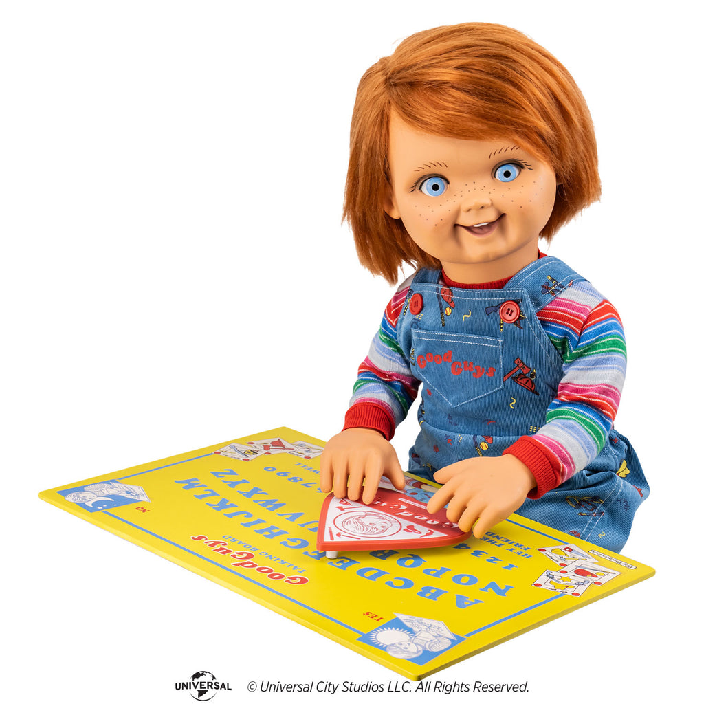 Good guys doll, blue eyes, red hair freckles, wearing blue overalls and striped shirt. Sitting in front of the yellow talking board, hands on the red and white planchette.