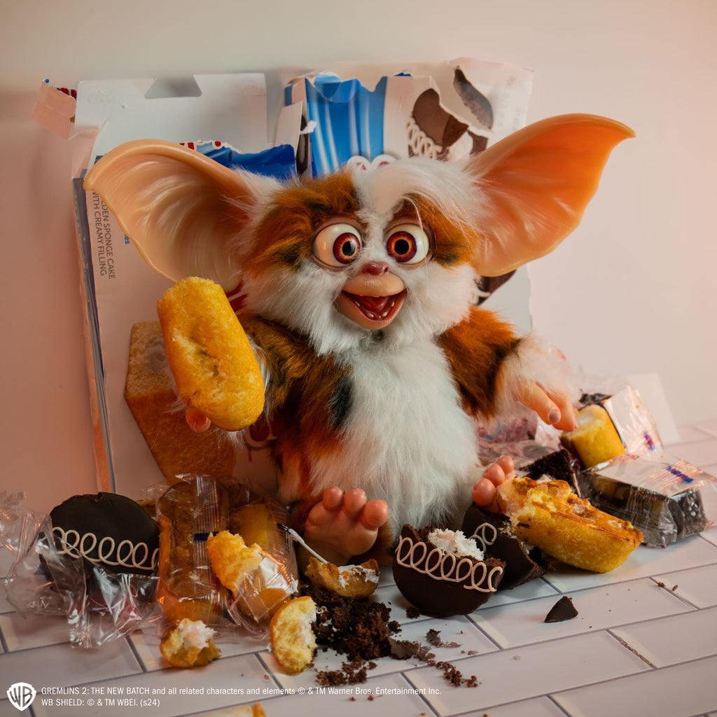 Gremlins 2 prop - Mogwai Daffy, brown and white with some black fur, large crossed eyes, mouth open in a smile, large pointed tan ears. Hairless hands and feet. sitting on a tiled counter surrounded by half-eaten snack cakes, holding one in his right hand
