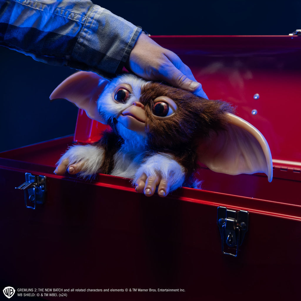 Glam shot.  Gizmo mogwai prop, sitting in a red toolbox, man's hand patting his head,  brown and white fur large brown eyes, large pointed tan ears. Hairless hands. White text at bottom is licensing information for the product.