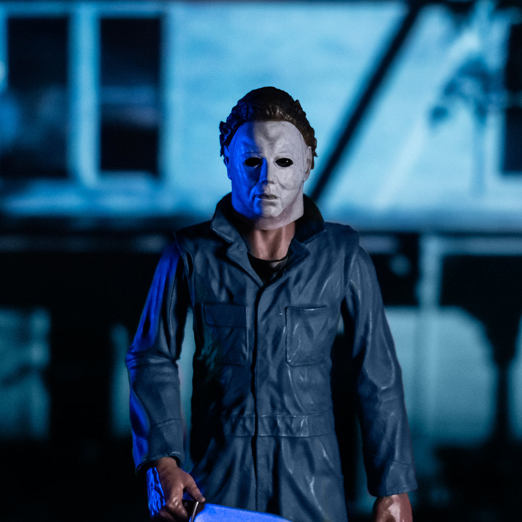 Glamour background, model house with nighttime lighting. closeup view in foreground Michael Myers 8 inch action figure shown from hips up. Wearing Halloween (1978) Michael Myers mask, dark coveralls. holding a kitchen knife in right hand.