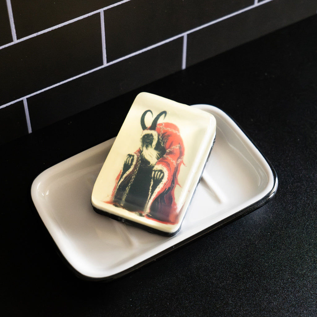 Bar soap. White, with illustration of Krampus, creature in red hooded robe with white trim, draped with chains, black face with mouth open, long curved black horns on head, clawed hands coming out of sleeves, under clear soap layer. Shown in white soap dish on black counter.