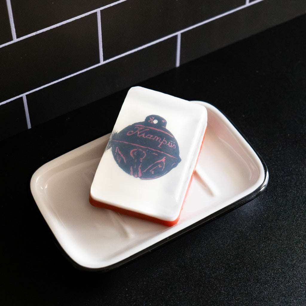 Bar soap. White, with illustration of black sleigh bell with embellishments, cursive text reads Krampus, under clear soap layer. Shown in white soap dish on black countertop