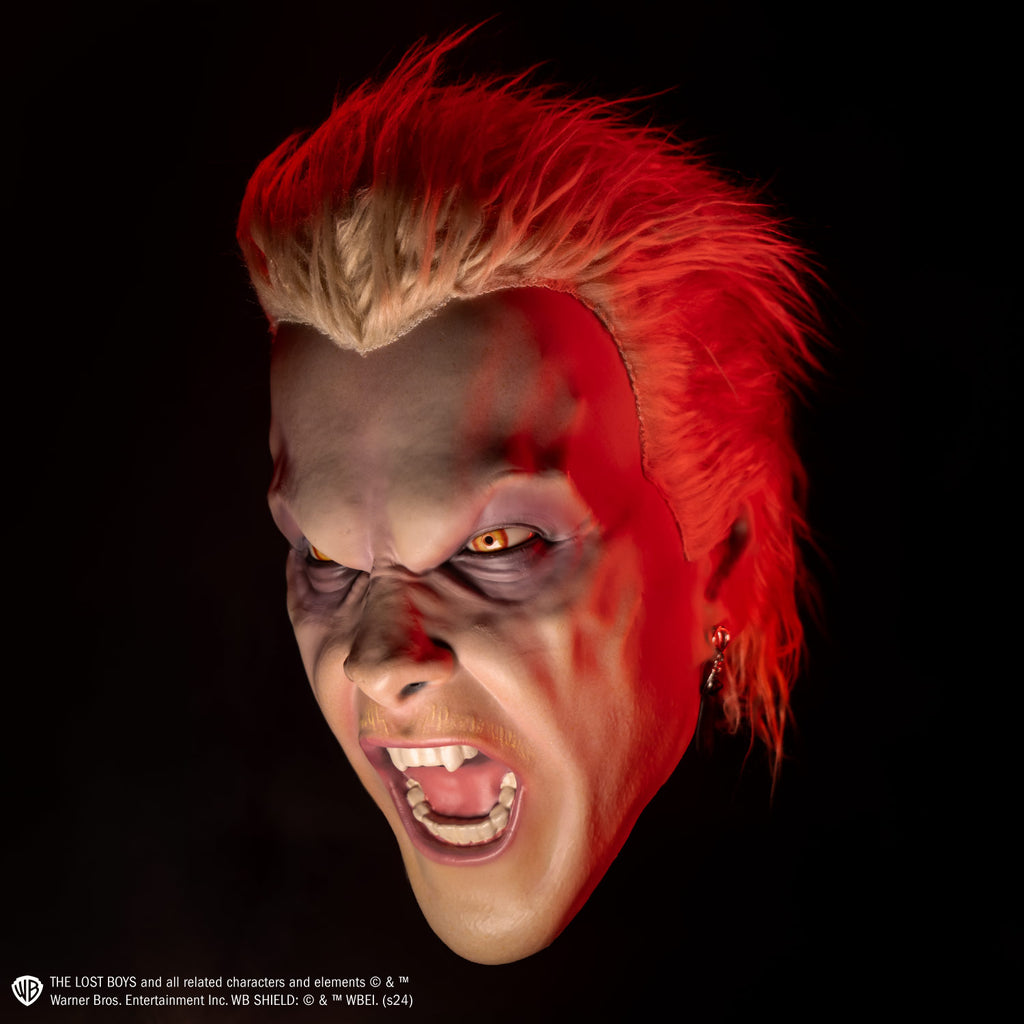 Mask, left view.  Light blond hair, slight mullet.  Furrowed brow, no eyebrows, dark circles under yellow eyes, long black earring in left ear, pale thin blond mustache,  mouth open in a snarl showing pink tongue and white teeth, fangs on either side of top two center teeth