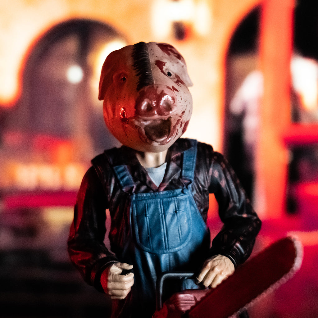 Orange Motel Hell glamour background. 8 inch figure closeup front view, head and torso to hips. Person in foreground with bloodied pig head mask, mouth open. Wearing flannel print shirt, blue overalls. Holding red chainsaw with left hand.