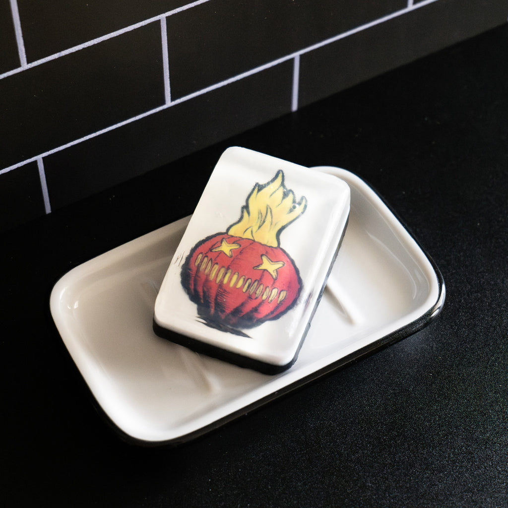 Bar soap. White, with illustration of Trick 'r Treat Sam-o-lantern under clear soap layer. Orange jack o' lantern face, two X eyes, several vertical hashmarks for a mouth, flames rising out of the top.  Shown in white soap dish on black counter.