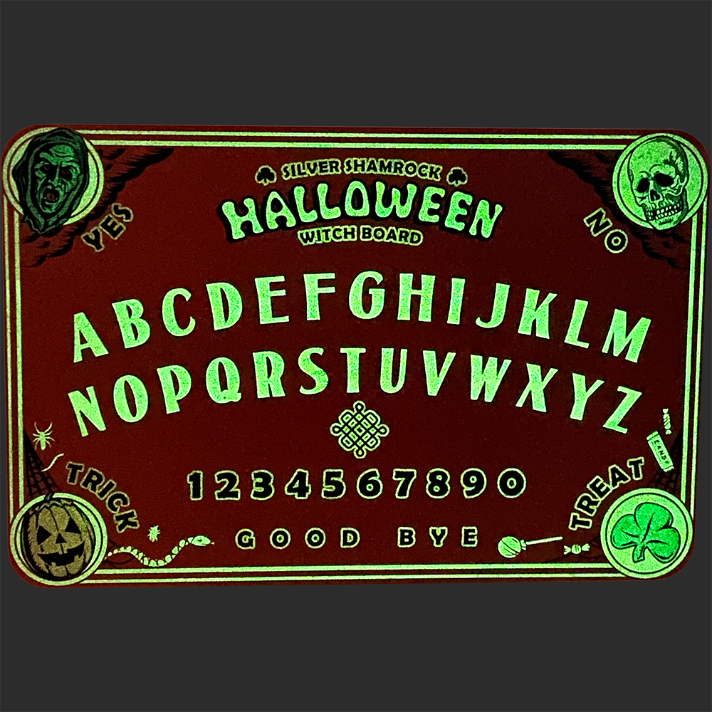 Witch board Glow feature. Top left corner green witch face, text reads Yes. Top right corner skull face, text reads no. Bottom left Jack o' lantern, text reads Trick. Bottom right corner, green shamrock, text reads Treat. Center of board, top to bottom text reads Silver Shamrock Halloween Witch Board. entire alphabet a through m and then N through Z, numbers 1 through 0 text at bottom reads Good Bye. 
