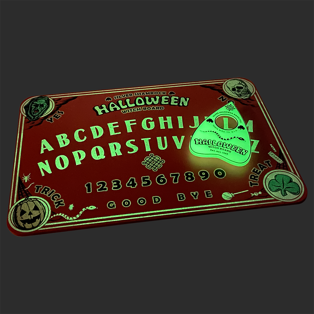 Witch board Glow feature. Top left corner green witch face, text reads Yes. Top right corner skull face, text reads no. Bottom left Jack o' lantern, text reads Trick. Bottom right corner, green shamrock, text reads Treat. Center of board, top to bottom text reads Silver Shamrock Halloween Witch Board. entire alphabet a through m and then N through Z, numbers 1 through 0 text at bottom reads Good Bye. Green glow planchette sitting on board.