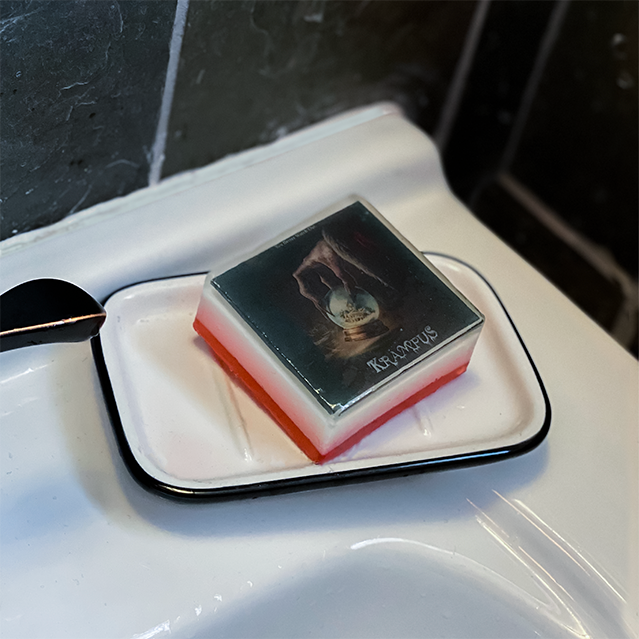 Bar soap. White, with illustration of a gnarled hand holding a snowglobe, white text reads Krampus, under clear soap layer. Shown in white and black soap dish on white sink