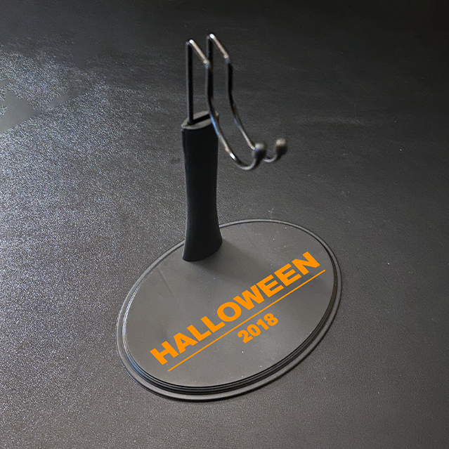 Figure stand for 12" action figure. Gray base, orange printed text reads Halloween 2018. Post with saddle hook to hold figure.
