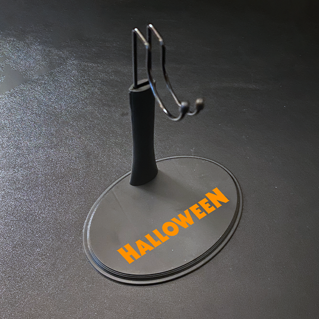 Figure stand for 12" action figure. Gray base, orange printed text reads Halloween. Post with saddle hook to hold figure.  