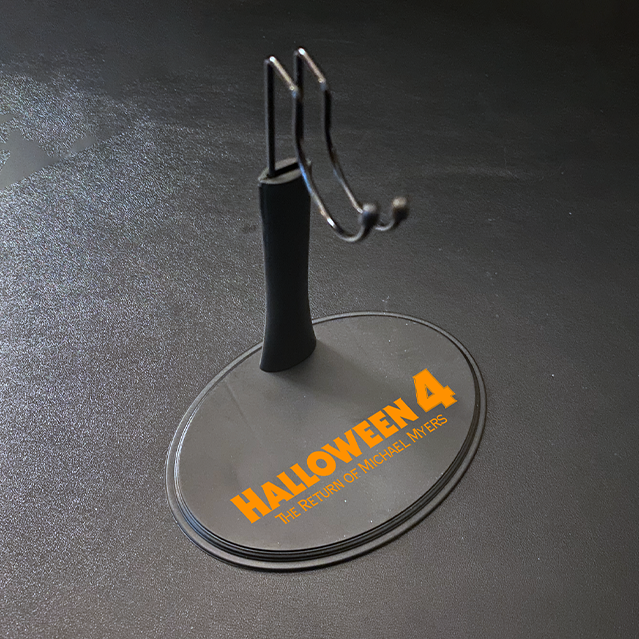 Figure stand for 12" action figure. Gray base, orange printed text reads Halloween 4 The Return of Michael Myers. Post with saddle hook to hold figure.