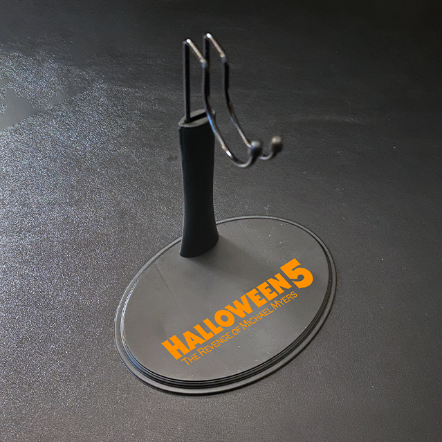 Figure stand for 12" action figure. Gray base, orange printed text reads Halloween 5 The Revenge of Michael Myers. Post with saddle hook to hold figure.