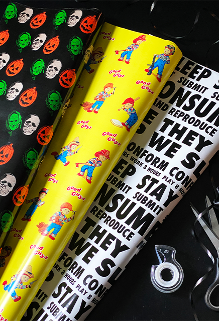 Three rolls of wrapping paper, black with witch, skull and pumpkin designs, Yellow with good guys designs, white with black text. Next to paper is ribbon, tape and scissors.