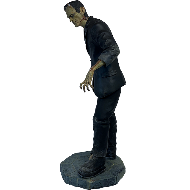 Frankenstein statue, front view. Pale greenish skin short black hair, metal bracket on top left forehead, wound on left jawline , heavy brow, dark circles around eyes, dark lips, metal bolts on sides of neck. Dirty black suit and pants, large black shoes, set on gray, stone textured base, skull between feet.