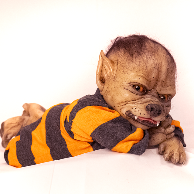 Doll. Full body view, lying on stomach, head resting on arms.  Tan skin, pointed ears on side of head, sparse black fur on head and arms. Gold eyes, canine nose, mouth with 2 fangs, pink tongue slightly sticking out. Paw like hands with claws. Wearing gray and orange striped pajamas.