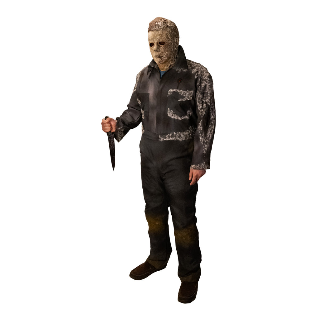 Man in Halloween Ends mask.  Wearing dark coveralls, dirty distressed and appearing to be covered in mold.  Holding knife in right hand.