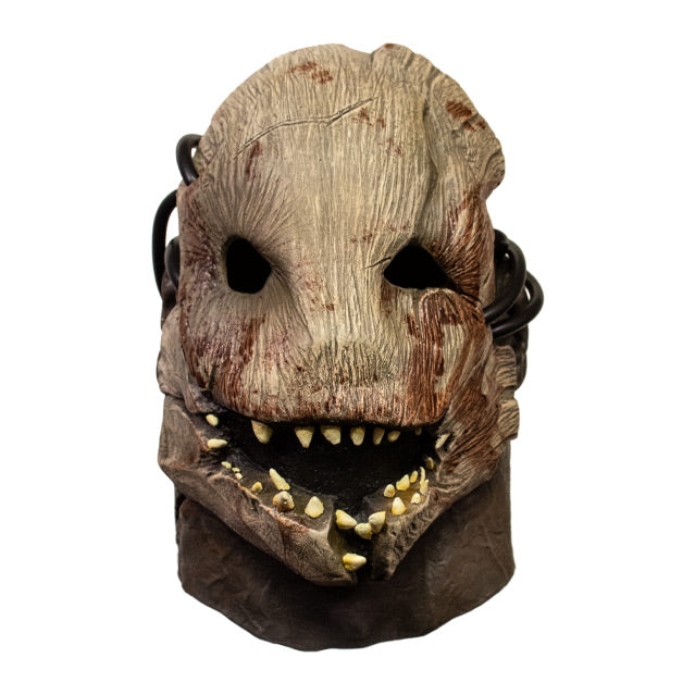 Front view. Trapper mask.  Woody creature facemask, brown with blood stains, many misaligned teeth set in crooked jaw, empty black eye sockets.  Attached with rubber tubing to human face, neck and ears showing.