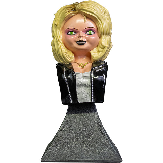 Front view. Tiffany mini bust, blond hair, green eyes. Black eyeliner, pink eye shadow, Black lipstick, black beauty mark on left above lip. Gold necklace cursive Tiff. Wearing a white wedding dress,  black leather jacket with silver zippers. Head, neck, and chest attached to gray stone textured base.