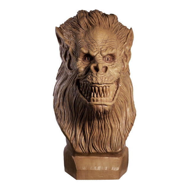 Front view. Crate Beast bust. Clay colored, beast face, wrinkled skin, eyes with vertical long pupils, pointed ears, snout nose, large grinning mouth with many sharp teeth, long hair covering head, lower face and neck.  Set on a base.