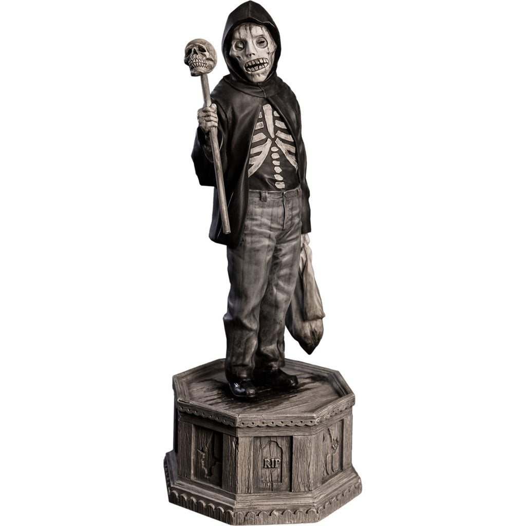 Front right view, statue.  Grayscale, Boy with skeletal face, wearing black hooded jacket over black skeleton shirt, jeans, black shoes. Holding baton topped with skull in right hand, holding dirty sack in left hand.  Standing on hexagon base made of gravestones