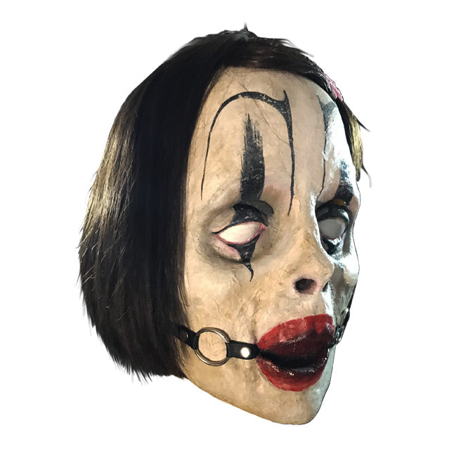right side view. Mask with dark brown hair with barrette, exaggerated black eyebrows and clown eye makeup, red lips, ball gag in mouth with straps