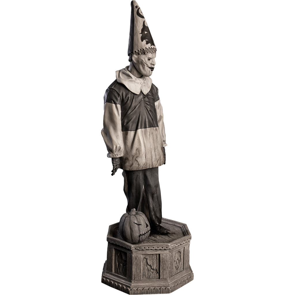 Front right view, statue. Grayscale, Creepy clown, tall pointy hat, black and white shirt with large white collar, black pants, shoes and gloves. Standing next to jack o' lantern on hexagon base made of gravestones