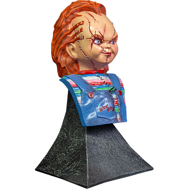 Right side view. Scarred Chucky mini bust, red hair, blue eyes, scarred face. Wearing a red, white, blue and green striped shirt, ripped blue overalls with red buttons, red Good Guys printed on pocket. Head, neck, and chest attached to gray stone textured base.