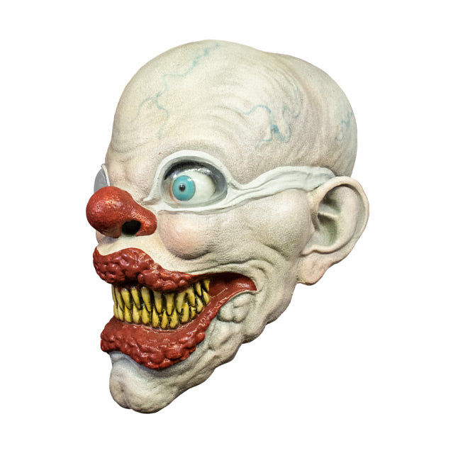 Left side view. Creepy bald clown face. White skin with blue veins. Light blue bulbous eyes, wearing white goggles. Large red nose. Enlarged, bumpy red lips, smiling. Many sharp yellow teeth.
