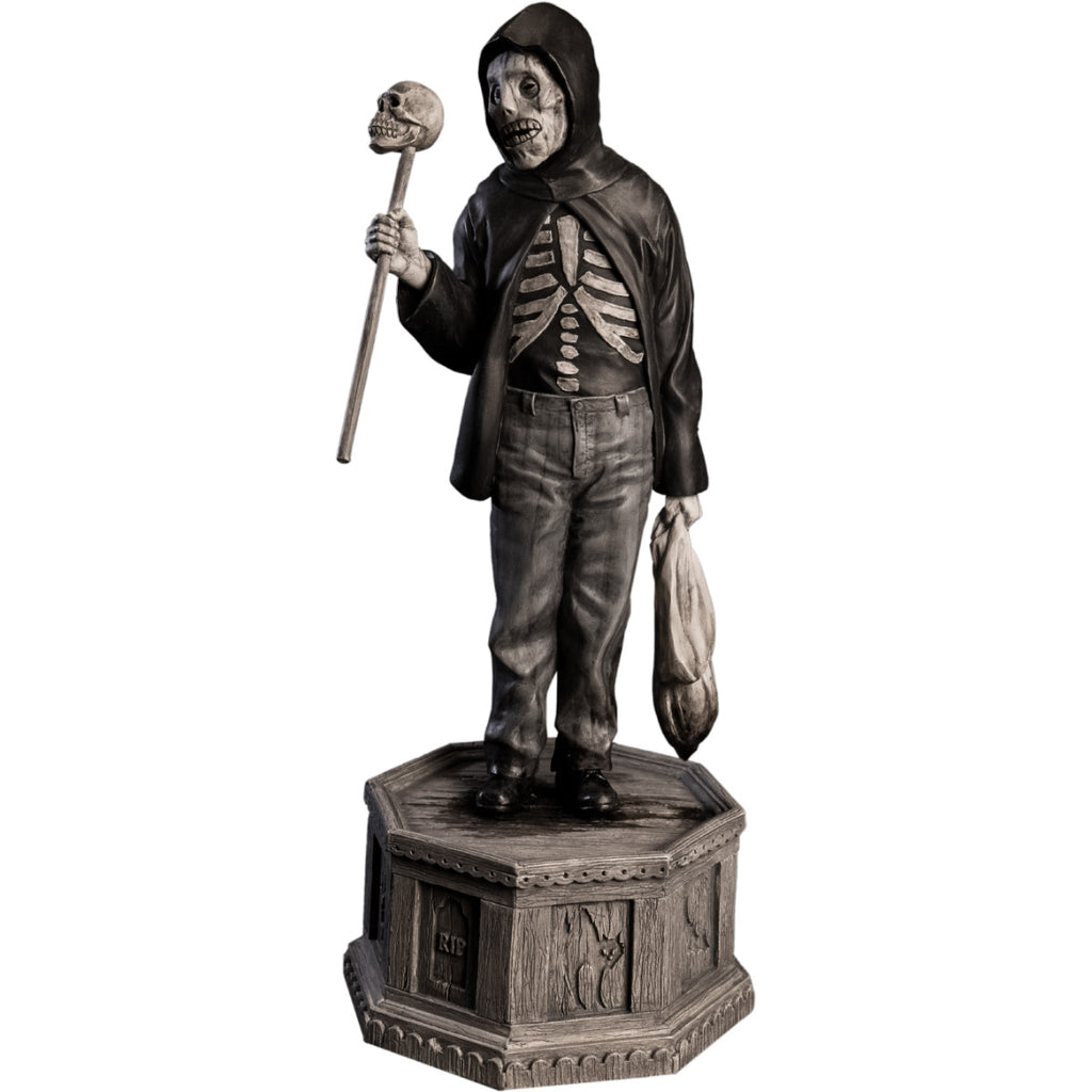 Front view, statue. Grayscale, Boy with skeletal face, wearing black hooded jacket over black skeleton shirt, jeans, black shoes. Holding baton topped with skull in right hand, holding dirty sack in left hand. Standing on hexagon base made of gravestones