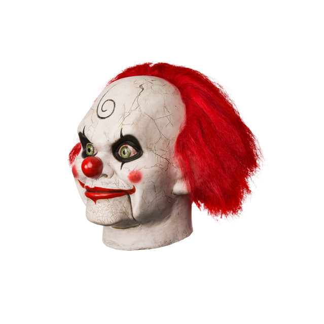 Left side view. Mary Shaw Clown Mask, head and neck. Distressed finish white skin, ventriloquist dummy clown face, spiral curl drawn on forehead, rosy red spots on cheeks, red nose, bright red hair, black-rimmed, bloodshot green eyes. Bright red grinning lips, hinged lower jaw.