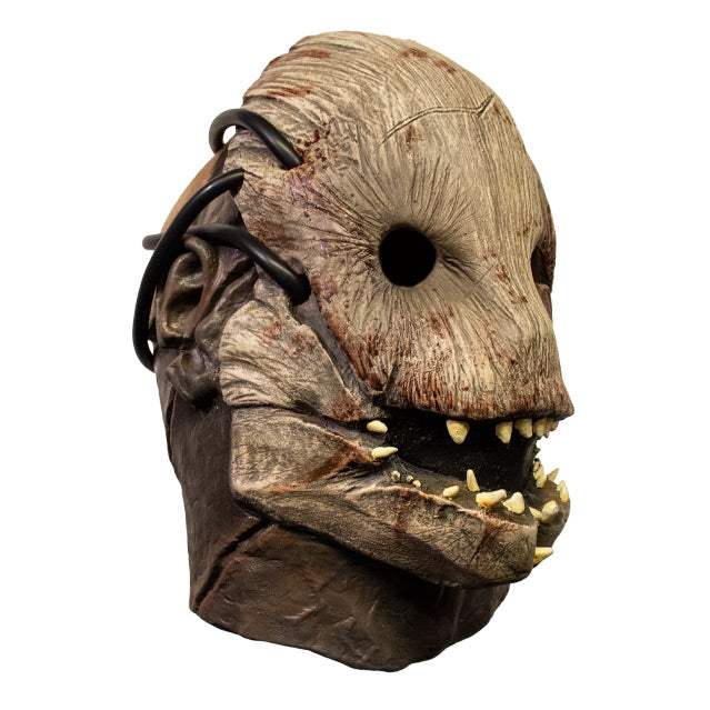 Right side view. Trapper mask. Woody creature facemask, brown with blood stains, many misaligned teeth set in crooked jaw, empty black eye sockets. Attached with rubber tubing to human face, neck and ears showing.