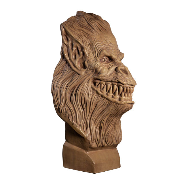 Right side view. Crate Beast bust. Clay colored, beast face, wrinkled skin, eyes with vertical long pupils, pointed ears, snout nose, large grinning mouth with many sharp teeth, long hair covering head, lower face and neck. Set on a base.