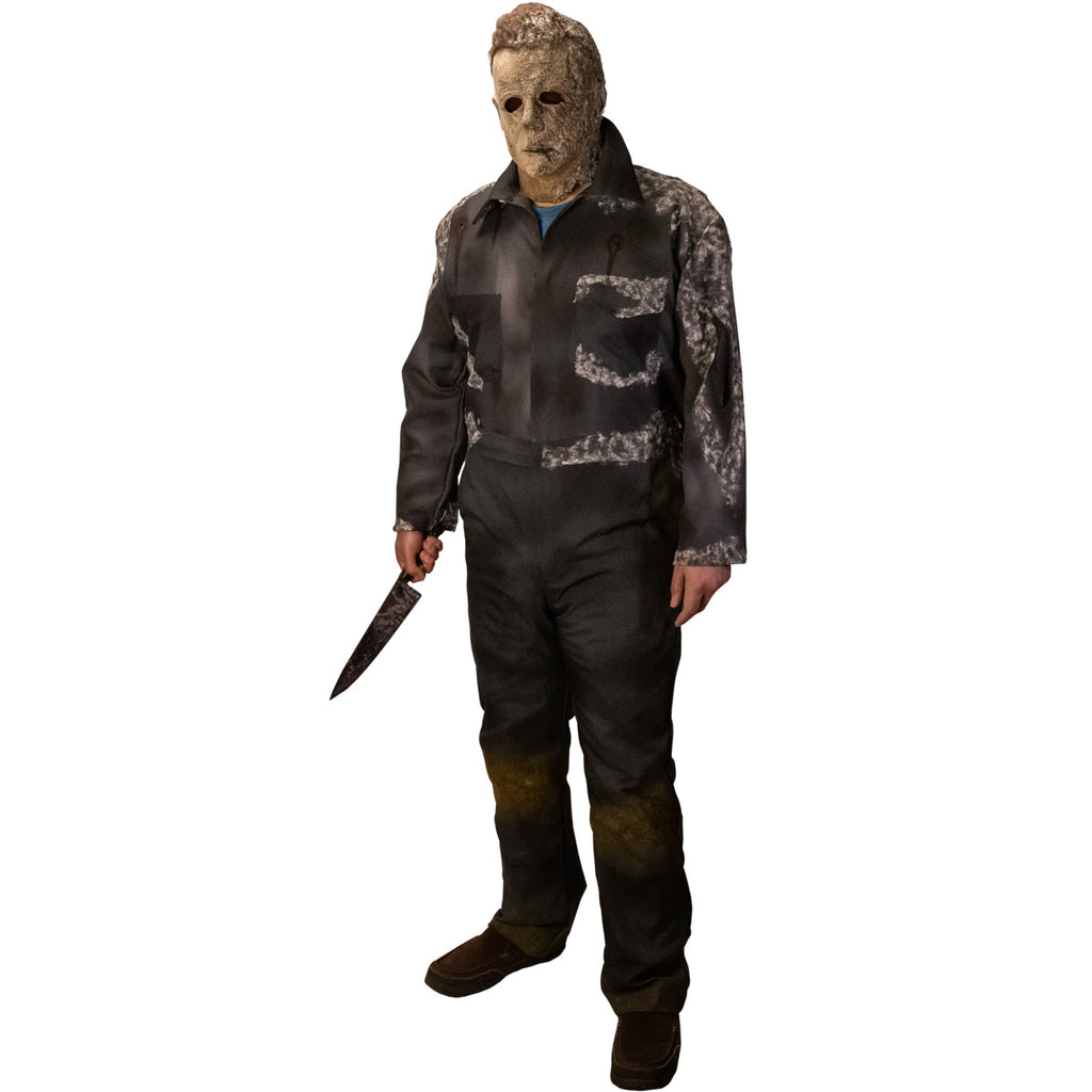 Man in Halloween Ends mask. Wearing dark coveralls, dirty distressed and appearing to be covered in mold. Holding knife in right hand.