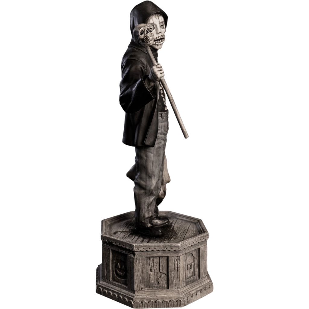 Side right view, statue. Grayscale, Boy with skeletal face, wearing black hooded jacket over black skeleton shirt, jeans, black shoes. Holding baton topped with skull in right hand, holding dirty sack in left hand. Standing on hexagon base made of gravestones
