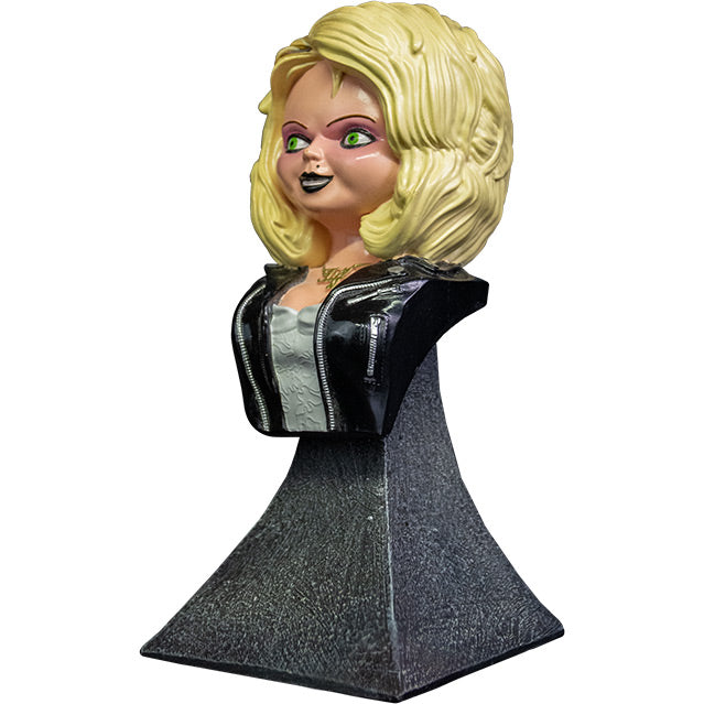 Left side view. Tiffany mini bust, blond hair, green eyes. Black eyeliner, pink eye shadow, Black lipstick, black beauty mark on left above lip. Gold necklace cursive Tiff. Wearing a white wedding dress, black leather jacket with silver zippers. Head, neck, and chest attached to gray stone textured base.