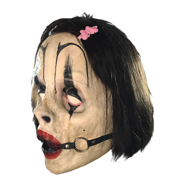 Left side view. Mask with dark brown hair with barrette, exaggerated black eyebrows and clown eye makeup, red lips, ball gag in mouth with straps
