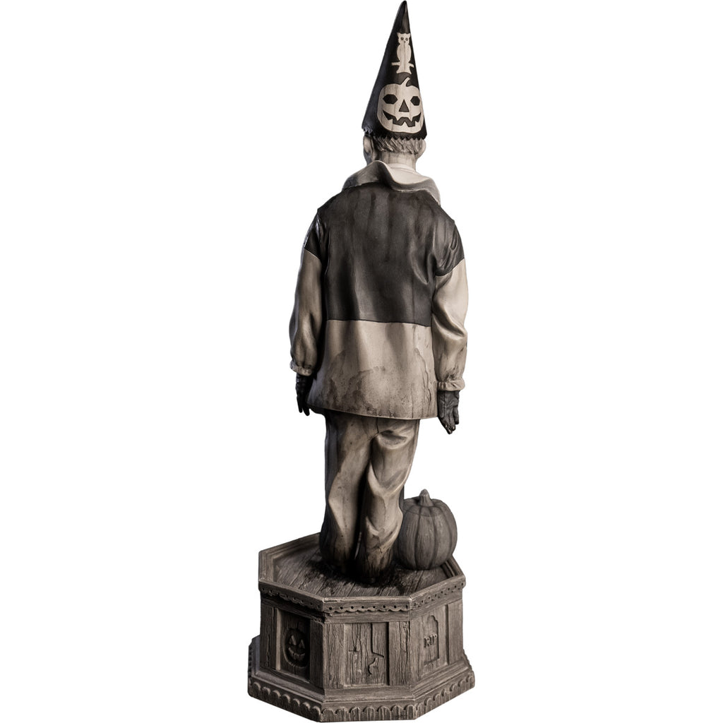 back view, statue. Grayscale, Creepy clown, tall pointy hat, black and white shirt with large white collar, black pants, shoes and gloves. Standing next to jack o' lantern on hexagon base made of gravestones