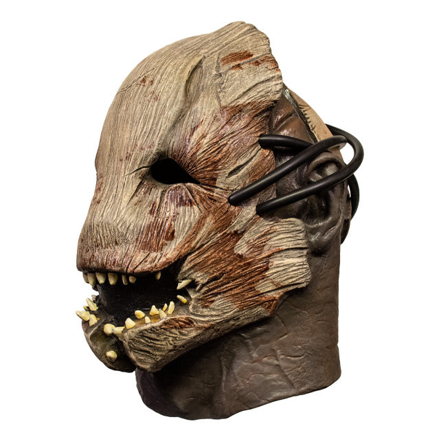 Left side view. Trapper mask. Woody creature facemask, brown with blood stains, many misaligned teeth set in crooked jaw, empty black eye sockets. Attached with rubber tubing to human face, neck and ears showing.