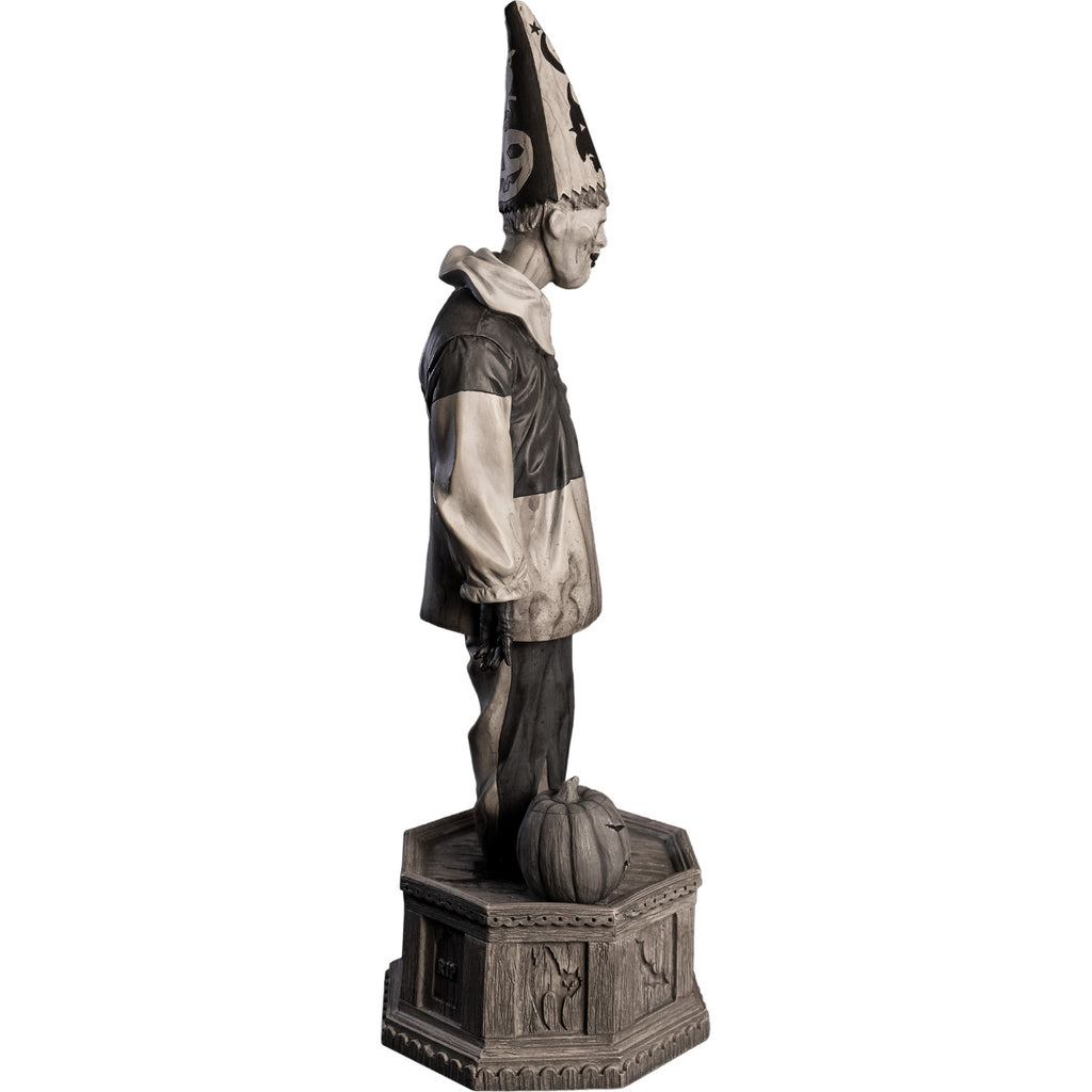 Right side view, statue. Grayscale, Creepy clown, tall pointy hat, black and white shirt with large white collar, black pants, shoes and gloves. Standing next to jack o' lantern on hexagon base made of gravestones