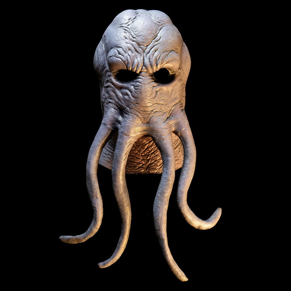 Mask, front view, head and neck, on black background with dramatic lighting. Lumpy octopus-like head, wrinkled gray flesh. Four long tentacles extending from middle of face.